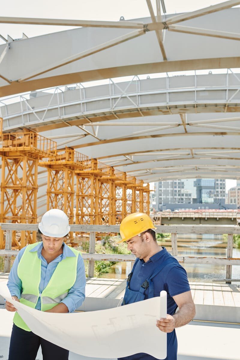 Fortschrittsbericht 5 Zwei Ingenieure auf einer Baustelle, die Baupläne mit Stahlkonstruktionen im Hintergrund prüfen.
